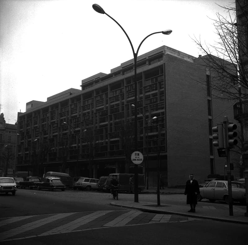 Grand immeuble moderne à cinq niveaux au coin d’une rue, voitures anciennes garées, photographie en noir et blanc