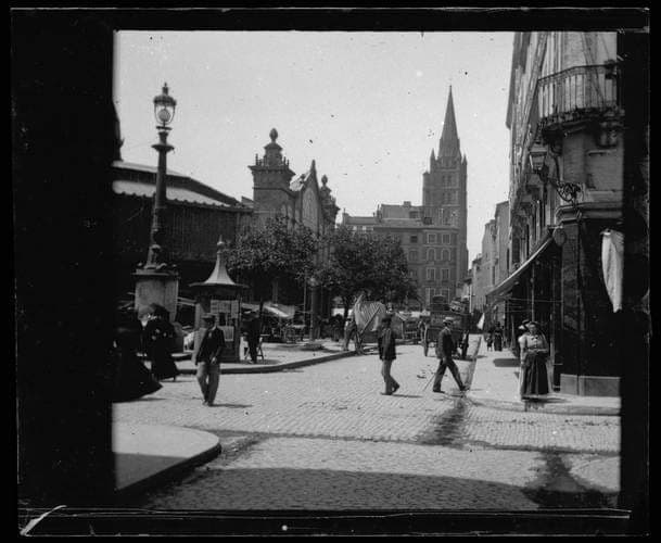 Place pavée avec la halle à gauche et un clocher au fond, passants en silhouettes, photographie ancienne