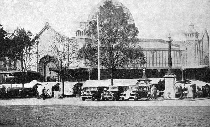 Photographie ancienne : halle de marché surmontée d’un dôme, étals bâchés et voitures devant