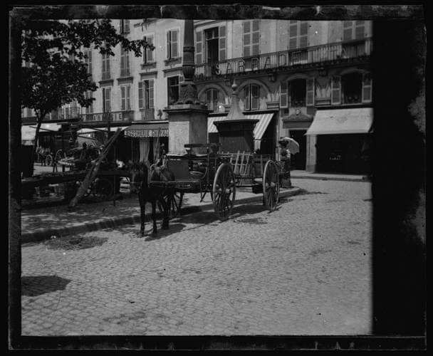 Photographie ancienne : charrette à cheval sur une place pavée devant des immeubles à balcons