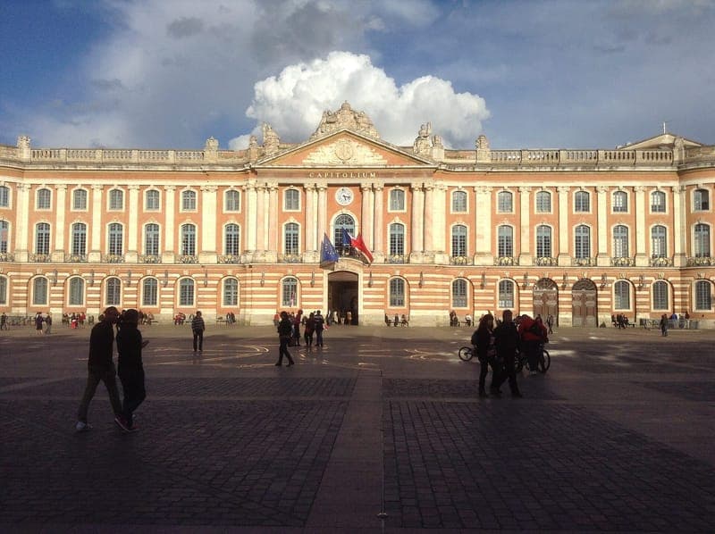 Façade du Capitole en brique rose et pierre sous un ciel nuageux, passants sur la place