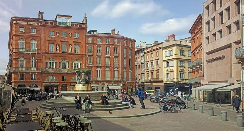Place de la Trinité : place pavée avec fontaine, terrasses et immeubles de brique, passants