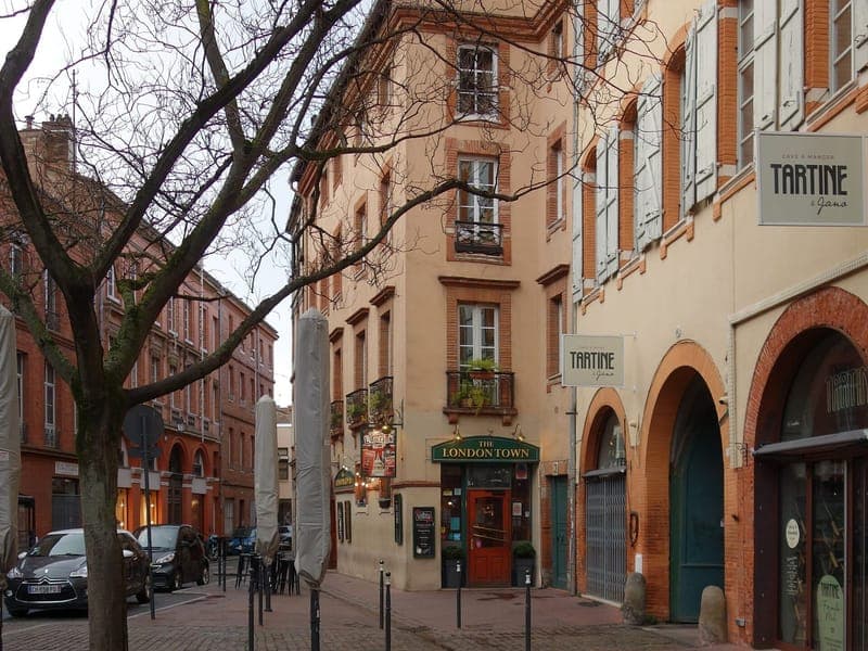 Angle de la place des Carmes avec façades de brique et d'enduit clair, terrasse de café et arbre sans feuilles