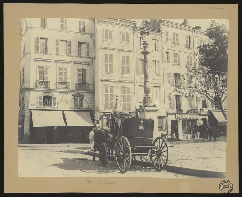 Calèche devant des immeubles à volets, avec un lampadaire au centre de la place