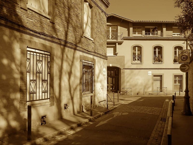 Rue Arnaud Baric : ruelle ensoleillée en teinte sépia, façades à grilles de fer forgé et arche au fond