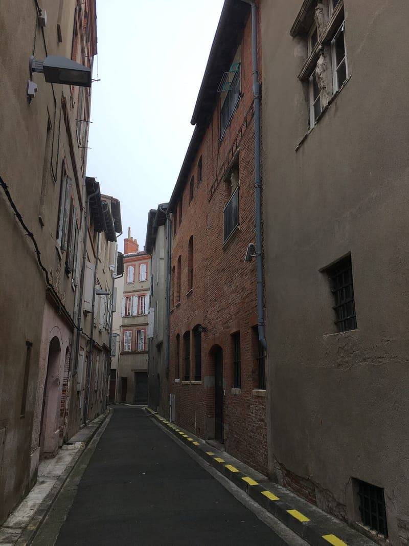 Rue de la Hache : rue très étroite entre des façades de brique et d'enduit, bandes jaunes au sol, immeuble rose au fond