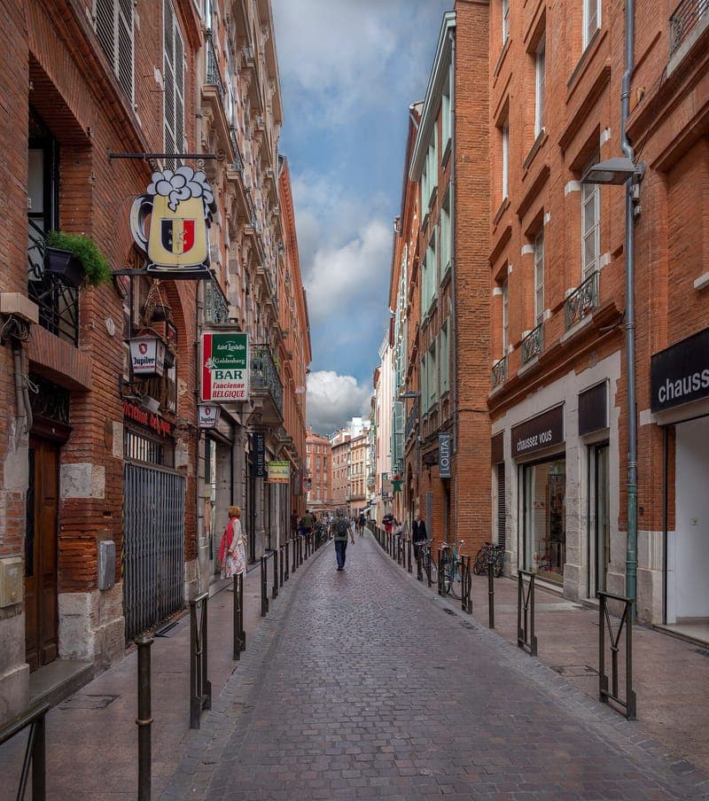 Rue de la Trinité : rue pavée étroite bordée de façades de brique, enseignes de commerces, passants lointains