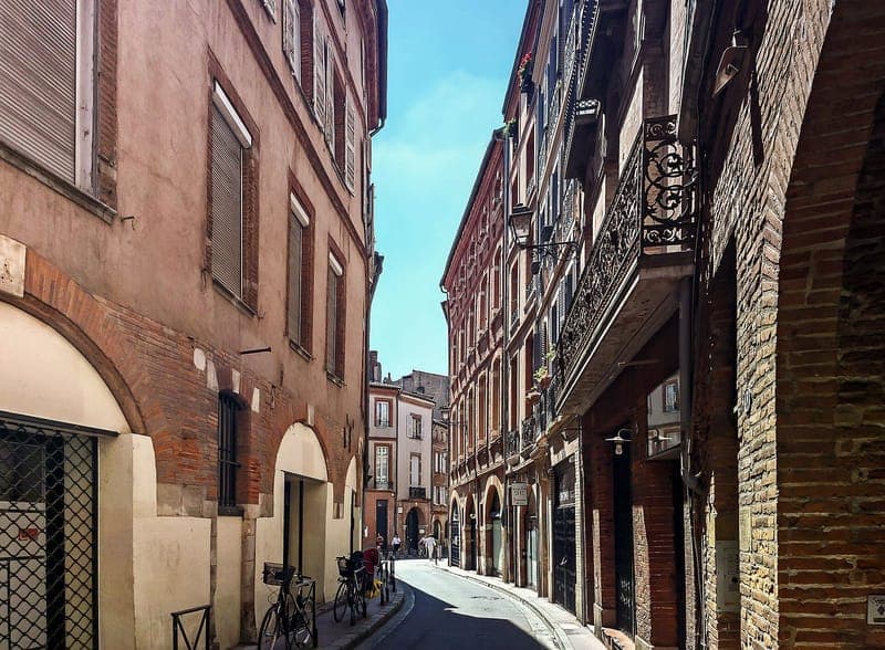 Rue des Paradoux : rue étroite bordée de façades de brique, balcons de fer forgé et arcades, ciel bleu au fond