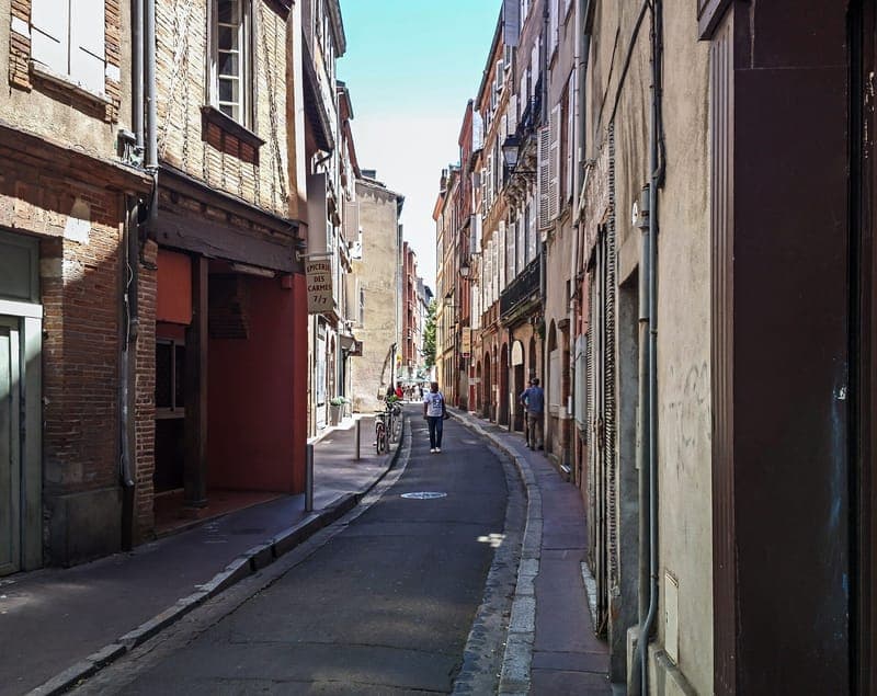 Rue étroite en perspective bordée de façades de brique, deux passants au loin
