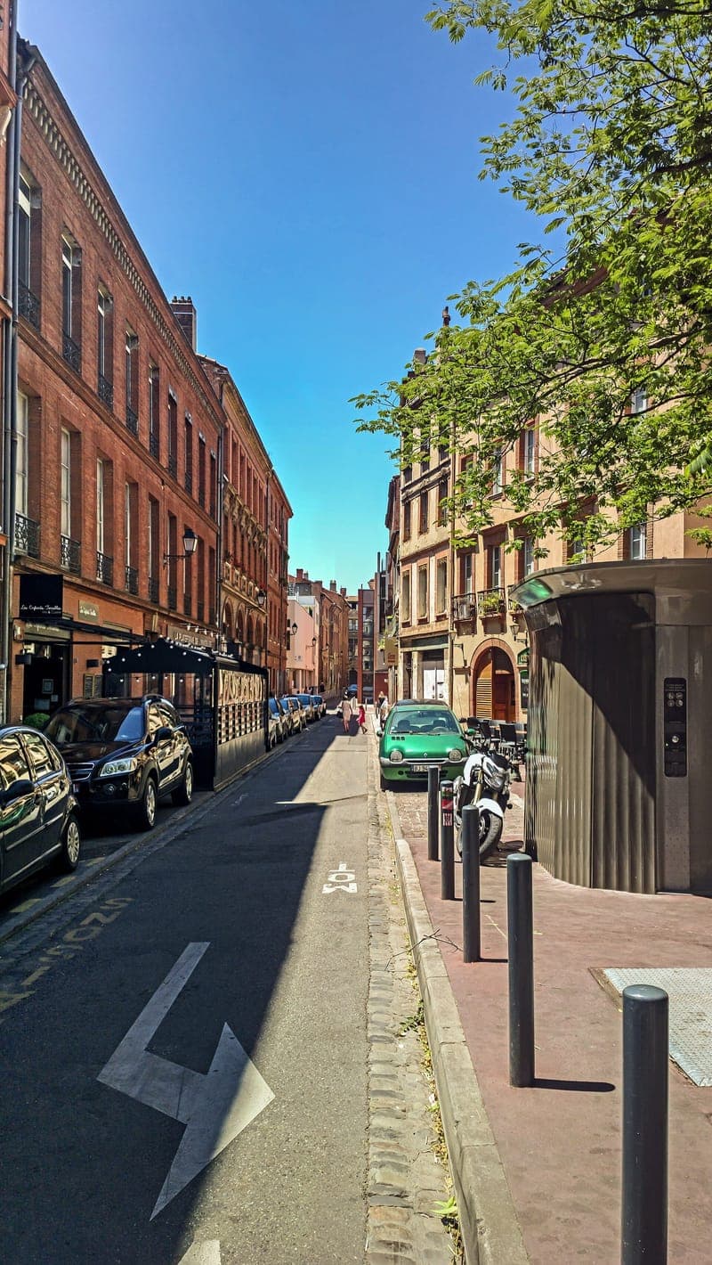 Rue des Prêtres : rue étroite bordée de façades de brique, voitures garées à droite, ciel bleu et arbre