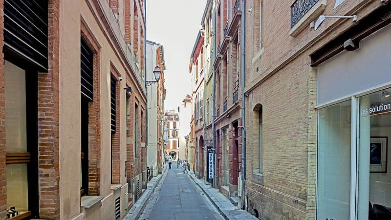 Rue étroite bordée de façades de brique et d'enduit, lanterne en fer forgé, passants au loin