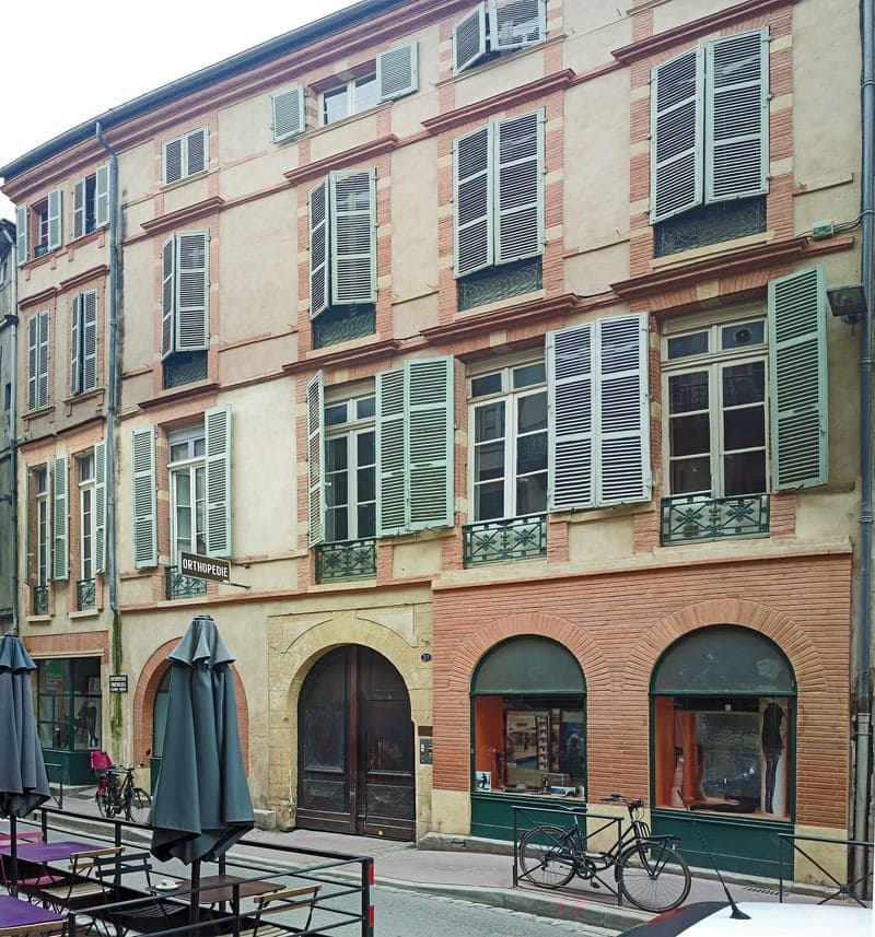 Façade de brique et de crépi à volets verts, rez-de-chaussée à arcades, terrasse de café au premier plan