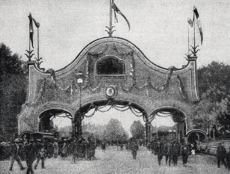 Photographie ancienne d’un arc de triomphe décoré de guirlandes et de drapeaux, foule lointaine en dessous