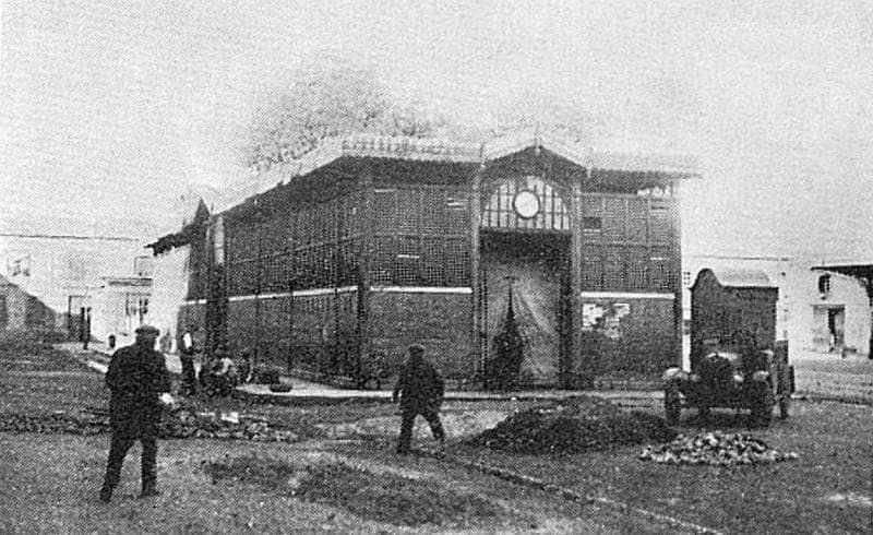 Photographie ancienne d’un marché couvert en brique, camionnette à droite, deux hommes au premier plan