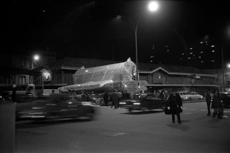 Photographie de nuit en noir et blanc : tronçon d’avion sous bâche devant un marché, voitures en mouvement