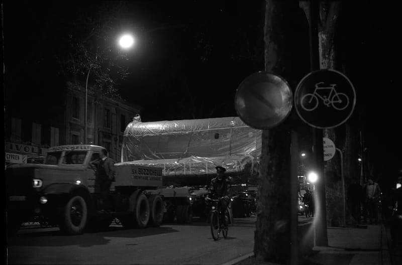 Photographie de nuit en noir et blanc : camion transportant un grand tronçon d’avion sous bâche, rue éclairée